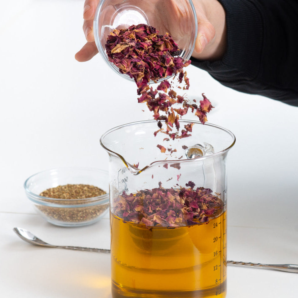 Person adding dried herbs into a glass container with liquid on a white surface.