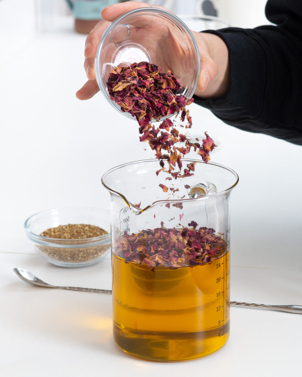 Person adding dried herbs into a glass container with liquid on a white surface.