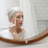 Woman applying skincare product in front of a mirror in a bathroom.
