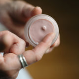 Hand holding a small container of pink cream against a blurred background