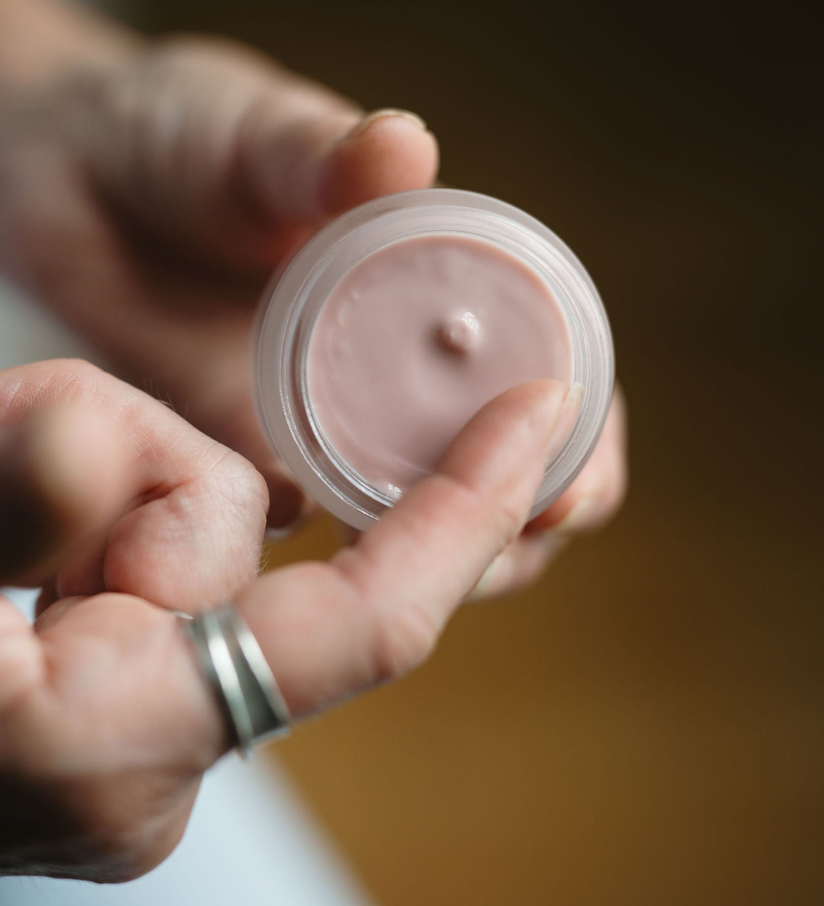 Hand holding a small container of pink cream against a blurred background