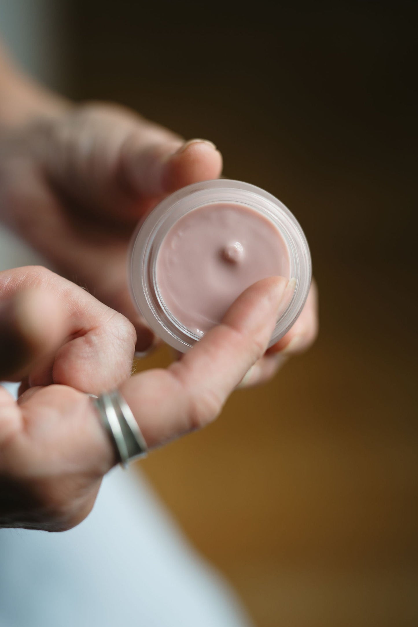 Hand holding a small container of pink cream against a blurred background