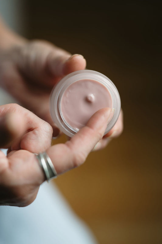 Hand holding a small container of pink cream against a blurred background